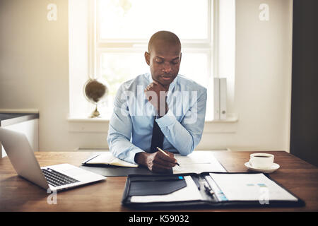 Junge afrikanische Executive sitzt an seinem Schreibtisch in einem Büro Lesen von Dokumenten und der an einem Notebook arbeitet Stockfoto