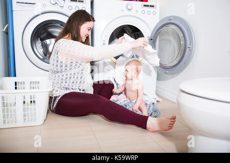 Mutter Hausfrau mit einem Baby in Wäscherei beschäftigt Falten Kleidung in die Waschmaschine Stockfoto