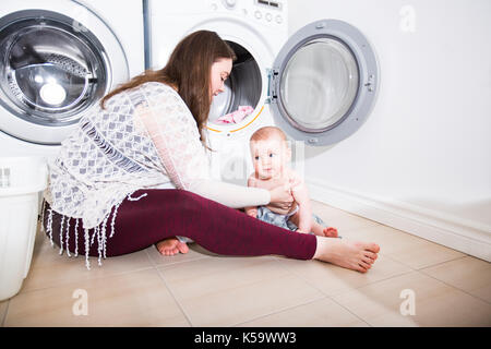 Mutter Hausfrau mit einem Baby in Wäscherei beschäftigt Falten Kleidung in die Waschmaschine Stockfoto