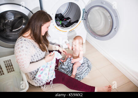 Mutter Hausfrau mit einem Baby in Wäscherei beschäftigt Falten Kleidung in die Waschmaschine Stockfoto