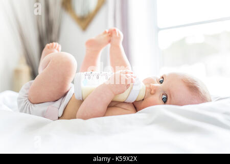 Hübsches Babymädchen trinkt Wasser aus der Flasche auf Bett liegend. Kind weared Windel im Kinderzimmer. Stockfoto