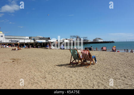 Leute sitzen auf Liegestühle auf Clacton-on-Sea Strand an einem sonnigen Tag Stockfoto