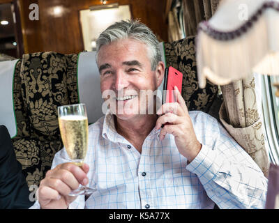 LUXUS LIFESTYLE-ZUG ERSTKLASSIGER CHAMPAGNER entspannter glücklicher reifer Mann, der auf seinem Smartphone sprach und seine Reise in der Pullman-Speiseeisenbahn genoss Stockfoto