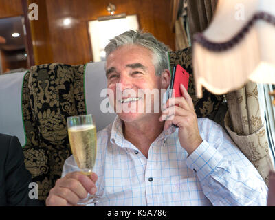 Entspannt reifer Mann mit einem Glas Champagner, Reden auf seinem iPhone 7 Plus, genießen eine Fahrt in einem Luxus vintage Pullman Waggon Stockfoto