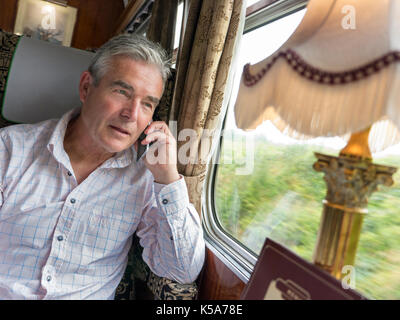 Entspannt reifer Mann in der First Class Restaurant Rampe Auto sprechen auf seinem iPhone 6, genießen eine Fahrt in einem Luxus vintage Pullman Waggon Stockfoto