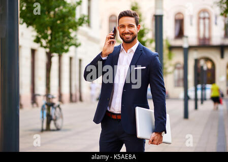 Ein hübscher junger Geschäftsmann mit seinem Laptop während Gespräch auf seinem Handy und stehen auf der Straße Stockfoto