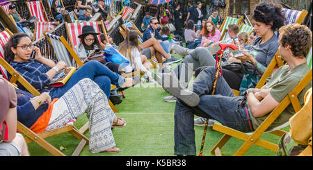 Die Leute sitzen und in Liegestühlen im Camden Market entspannen, Die Stühle sind auf Kunstrasen auf verschiedenen Ebenen eines Farbverlaufs einstellen. Stockfoto