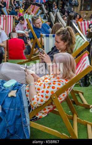 Die Leute sitzen und in Liegestühlen im Camden Market entspannen, Die Stühle sind auf Kunstrasen auf verschiedenen Ebenen eines Farbverlaufs einstellen. Stockfoto