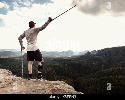 Happy Tourist mit unterarm Krücke über den Kopf erreicht Berggipfel. Wanderer mit verletzten Knie in "Wegfahrsperre und Medizin Polen halten Sie die Hand in die Luft. Colorfu Stockfoto