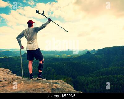 Happy Tourist mit unterarm Krücke über den Kopf erreicht Berggipfel. Wanderer mit verletzten Knie in "Wegfahrsperre und Medizin Polen halten Sie die Hand in die Luft. Colorfu Stockfoto