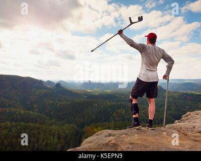 Happy Tourist mit unterarm Krücke über den Kopf erreicht Berggipfel. Wanderer mit verletzten Knie in "Wegfahrsperre und Medizin Polen halten Sie die Hand in die Luft. Colorfu Stockfoto