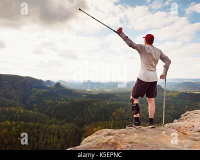 Happy Tourist mit unterarm Krücke über den Kopf erreicht Berggipfel. Wanderer mit verletzten Knie in "Wegfahrsperre und Medizin Polen halten Sie die Hand in die Luft. Colorfu Stockfoto