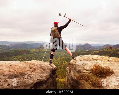 Touristische mit Medizin Krücke über den Kopf erreicht Berggipfel. Wanderer mit gebrochenen Bein in der Wegfahrsperre. Tiefe nebligen Tal unten Silhouette der glücklichen Mann Stockfoto