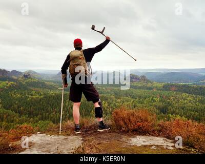 Wanderer mit gebrochenen Bein in der Wegfahrsperre. Touristische mit Medizin Krücke über den Kopf erreicht Berggipfel. Tiefe nebligen Tal unten Silhouette der glücklichen Mann Stockfoto