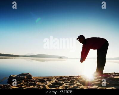 Tiefes Atmen runner Silhouette mit den Händen auf die Knie gebeugt. Seitenansicht zu Sport aktiven Mann stretching Muskeln und Bewegung am Meer Strand. Sun spiegeln Stockfoto