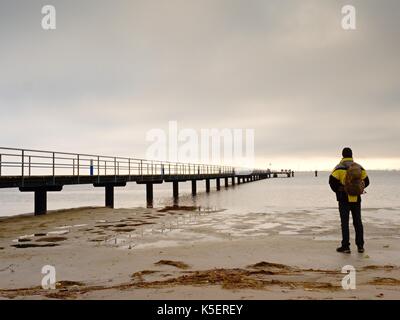 Mann Wanderer mit Rucksack stand alone auf schmutzigen Sandstrand und romantischen farbenfrohen Sonnenaufgang über hölzerne Seebrücke. Vintage Style getönten Wirkung Stockfoto