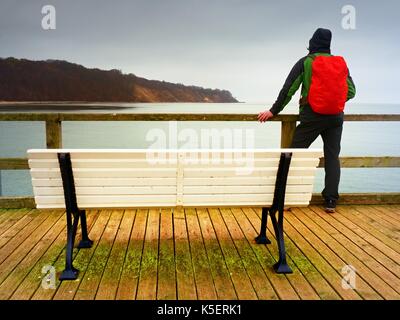 Touristische mit roten Rucksack auf hölzernen Pier über dem Meer. Mann in trekking Anzug im Hafen in dunklen regnerischen Tag. Touristische Mole, nasses Holz- Etage über dem Meer. Stockfoto