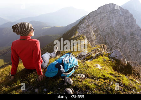 Frau in Rot wandern Jacke sitzen auf Gras auf dem Berg mit herrlicher Aussicht auf die Gipfel. Kladivo, Karakvanke, Slowenien. Stockfoto