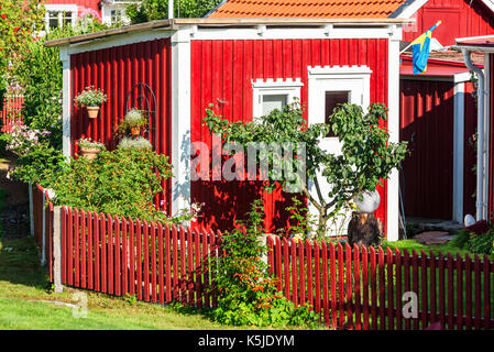 Rote und weiße Holz Gartenhaus mit viel Vegetation und Sträucher außerhalb. Red Lattenzaun im Vordergrund. Stockfoto