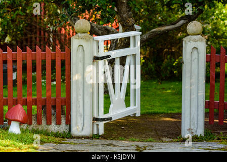 White Garden Gate auf rot Lattenzaun öffnen. Lane und Baum im Hintergrund. Stockfoto