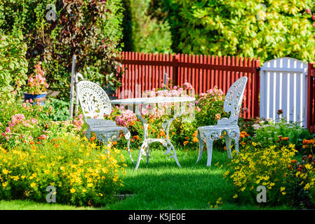 Weißes Metall Gartenmöbel unter den Blumen auf einer Wiese. Lattenzaun im Hintergrund. Stockfoto