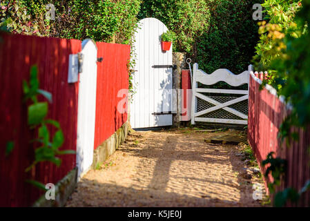 White Garden Gates am Ende einer Kies Trail. Red Lattenzaun Seiten der Fahrspur. Blumentopf am Tor. Stockfoto