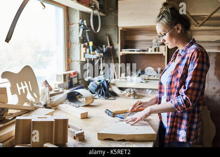 Profil anzeigen von selbstbewussten jungen Handwerkerin in Brillen Messung Holzbrett mit Bleistift und Lineal, während Tabelle steht in unordentlichen Workshop Stockfoto