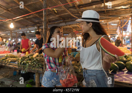 Zwei schöne Frauen Verkostung Wassermelone auf den traditionellen Markt in Asien jungen Mädchen Touristen kaufen frische Früchte auf Thailand Basar Stockfoto