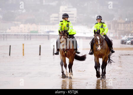 Zwei Bodenwanne Polizei Frau, auf Patrouille, in fluoreszierenden Uniformen reiten. ihre Polizei Pferde am Strand in nassen windigem Wetter. Stockfoto