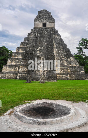 Ein hoch aufragender, antiker maya-Tempel steht vor einem Hintergrund von üppigen Bäumen unter einem bewölkten Himmel, mit einer alten kreisförmigen Struktur im Vordergrund Stockfoto