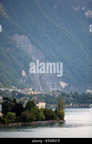 Chateau de Chillon am Genfer See in der Schweiz von üppigen Bäumen umgeben ist. Im Hintergrund sind riesige Berge an der alpinen Basis. Stockfoto