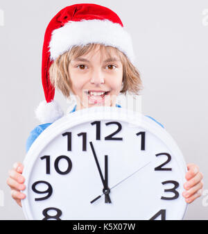 Emotionale Portrait von attraktiven kaukasische Mädchen tragen Santa Claus Red hat. Lustig nett lächelnd Kind bis 10 Jahr alt sind und große Uhr auf Grau backgr Stockfoto