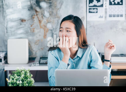 Jungen asiatischen casual Geschäftsfrau Gähnen vor Laptop Computer und Suchen über office Fenster mit Langeweile, harte Arbeit Konzept Stockfoto