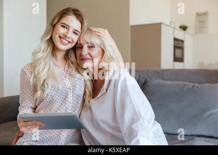 Bild der glücklichen jungen Dame zu Hause mit ihrer Großmutter auf Tablet Computer sitzen. Suchen Kamera. Stockfoto