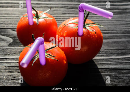 Drei reife rote Tomaten mit Strohhalmen auf schwarzen Holztisch Close-up. Frischer Saft gesundes Essen Konzept. Tomaten wie ein Gefäß mit frischem Saft Stockfoto