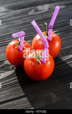 Drei reife rote Tomaten mit Strohhalmen auf schwarzen Holztisch Close-up. Frischer Saft gesundes Essen Konzept. Tomaten wie ein Gefäß mit frischem Saft Stockfoto