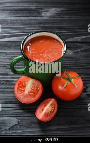 Tomatensaft in Grün Emaille Tasse auf einem schwarzen Holztisch unter Frische Tomaten Ansicht von Oben. Stockfoto