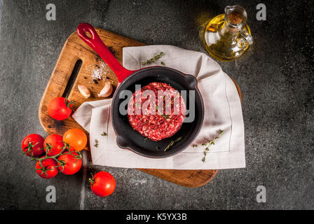 Raw organic Rindfleisch Burger Schnitzel mit Gewürzen, Thymian, Tomaten, Olivenöl in der Pfanne auf schwarzem Hintergrund, kopieren Raum Stockfoto