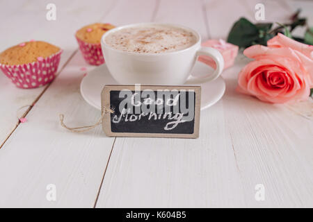 Kaffeebecher mit Kuchen, Rose und Noten guten Morgen auf weißem rustikalen Tisch von oben, leckeres Frühstück Stockfoto