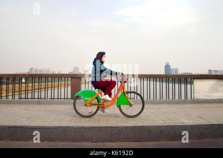 Stadt Taiyuan, Provinz Shanxi. Junge Frau mit Luftverschmutzung Maske Fahrten city Community bike teilen Zyklus über das Fen Fluss Stockfoto