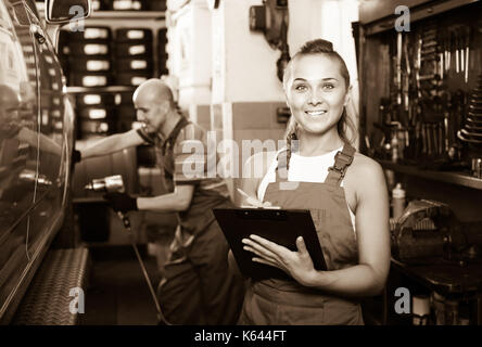 Freundlich lächelnden jungen Frau in Uniform stehen und schreiben Sie die Daten in auto Werkstatt Stockfoto