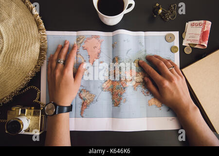 Frau, die Ihre Reise planen. Menschliche Hände auf der Weltkarte mit dem Hut, Kamera, etwas Geld, Notebook aus Recyclingpapier und frisch gebrühten Kaffee Stockfoto