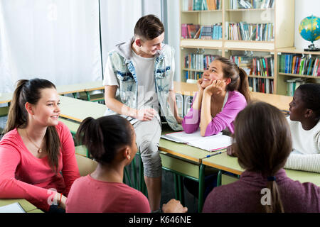 Glückliche junge Studenten verschiedener Nationalitäten sitzt und spricht im Klassenzimmer während der Pause Stockfoto