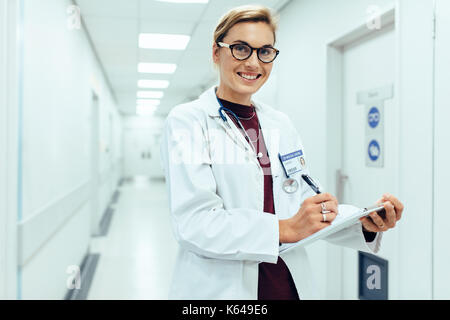 Portrait von lächelnden Frau Doktor im Krankenhaus Flur mit Zwischenablage. Kaukasische Medizin Praktiker schreiben medizinische Bericht und Ergebnisse Stockfoto