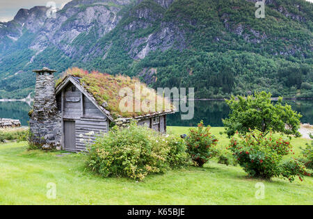 Alte märchenhafte Haus aus Holz mit Kamin aus gestapelten Schiefer und ein Dach mit Pflanzen und Bäume an einem Fjord in Norwegen mit Garten mit roten Blumen ein Stockfoto