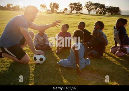 Grundschule Kinder und Lehrer sitzen mit Ball im Feld Stockfoto