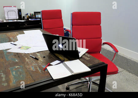 Zwei rote Bürostühle an einem Tisch sitzen mit einem Mac Book Laptop öffnen neben einem der Stapel Papier. Stockfoto