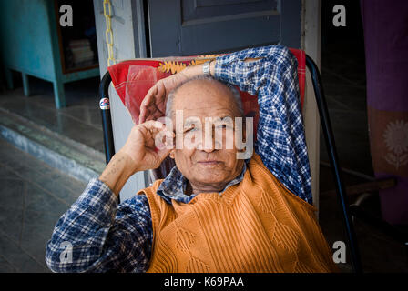 Porträt der alte Mann in einem Stuhl sitzend trägt ein T-Shirt und orange sleeveless Jumper in Nyaung Shwe, Inle Lake, Myanmar, Birma, Südostasien Stockfoto