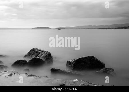 Eine minimalistische, Langzeitbelichtung Foto von einige große Felsen in einen See, mit perfekt noch Wasser und bewölkter Himmel Stockfoto