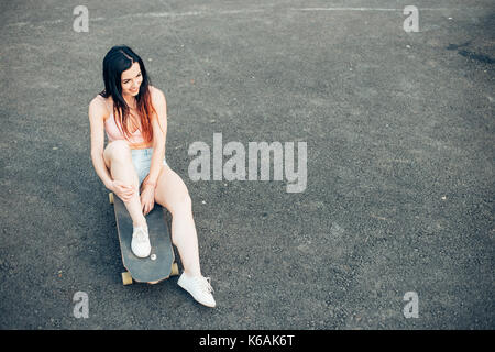 Schöne junge Mädchen mit Tätowierungen sitzt auf Longboard bei schönem Wetter Stockfoto